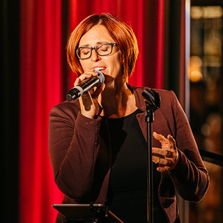 Sängerin mit roten, kurzen Haaren und Brille singt am Mikrofon.